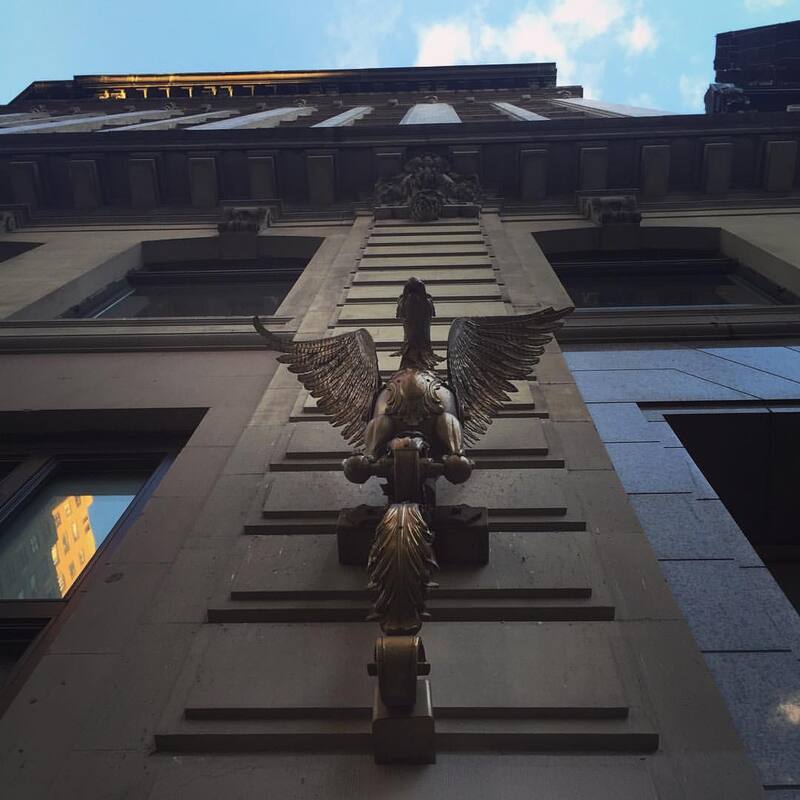 Upward image of a building with a large bird statue and blue sky and clouds reflecting on the building window.  New York City 2015.